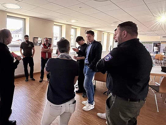 Eine kleine Gruppe von Erwachsenen steht in einem hellen Raum und diskutiert vor großen Plakaten an der Wand; eine Person erklärt etwas, während die anderen aufmerksam zuhören.