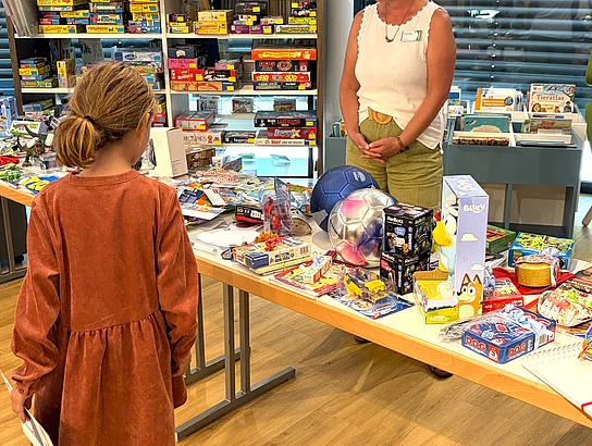 Ein Kind steht vor einem langen Tisch mit zahlreichen Preisen wie Spielen, Büchern, einem Fußball und weiteren Geschenken, während eine Mitarbeiterin des Büchereiteams dahinter wartet.