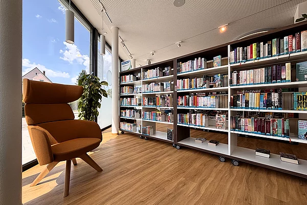 Ein gemütlicher Lesebereich mit orangefarbenem Sessel direkt an einer großen Fensterfront. Mehrere Bücherregale stehen auf Rollen und bieten zahlreiche Romane und Sachbücher. Tageslicht fällt durch die hohen Fenster und sorgt für eine ruhige Atmosphäre.