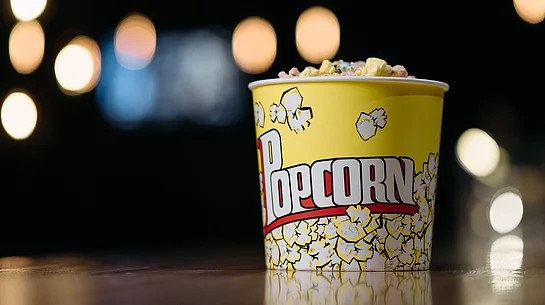 Ein gelber Popcornbecher, gefüllt mit buntem Popcorn, steht auf einer Oberfläche vor einem dunklen Hintergrund mit unscharfen, leuchtenden Lichtpunkten.