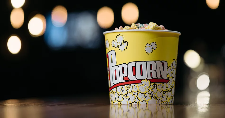 Ein gelber Popcornbecher, gefüllt mit buntem Popcorn, steht auf einer Oberfläche vor einem dunklen Hintergrund mit unscharfen, leuchtenden Lichtpunkten.