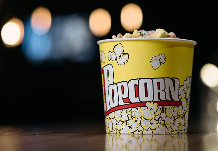 Ein gelber Popcornbecher, gefüllt mit buntem Popcorn, steht auf einer Oberfläche vor einem dunklen Hintergrund mit unscharfen, leuchtenden Lichtpunkten.