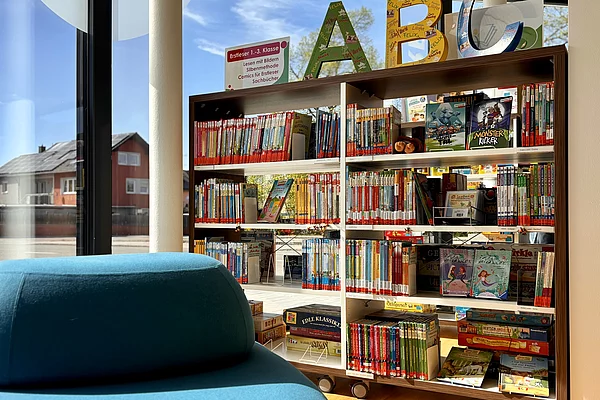 Ein heller Bereich der Stadtbücherei mit Kinder- und Jugendbüchern in niedrigen Regalen direkt an einer großen Fensterfront. Im Vordergrund befindet sich eine große, blaue Sitzgelegenheit. Oberhalb der Regale markieren bunte Buchstaben den Bereich. Tageslicht und offene Gestaltung schaffen eine freundliche Atmosphäre.