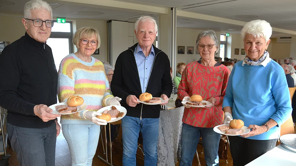 Fünf ältere Personen stehen in einem hellen Raum nebeneinander und halten jeweils einen Teller mit einem Brötchen und einer Scheibe Leberkäse, während im Hintergrund weitere Menschen an Tischen sitzen.