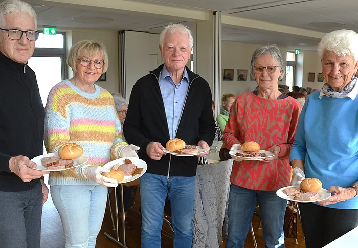 Fünf ältere Personen stehen in einem hellen Raum nebeneinander und halten jeweils einen Teller mit einem Brötchen und einer Scheibe Leberkäse, während im Hintergrund weitere Menschen an Tischen sitzen.