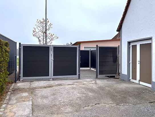 Ein gepflasterter Hof mit einem weißen Gebäude und geschlossenen dunkelgrauen Toren, dahinter ist ein kleiner Baum mit gelben Blättern und ein Flutlichtmast zu sehen.