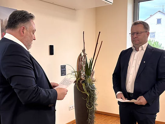 Ein Mann im Anzug liest aus einem Blatt Papier vor, während ein zweiter Mann ihm gegenübersteht und ebenfalls ein Dokument in den Händen hält. Die Szene wirkt offiziell und konzentriert – vermutlich ein Moment der Vereidigung. Im Hintergrund steht eine dekorative Pflanze, daneben fällt Tageslicht durch ein Fenster.