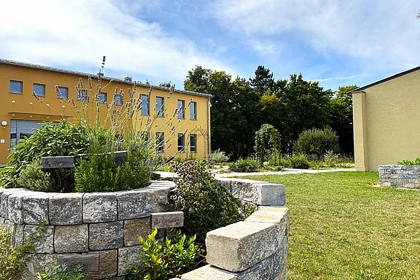 Im Vordergrund ein bepflanztes und gemauertes Kräuterbeet in Schneckenhausform. Im Hintergrund das orange-gelbe Gebäude des Mehrgenerationenhauses.