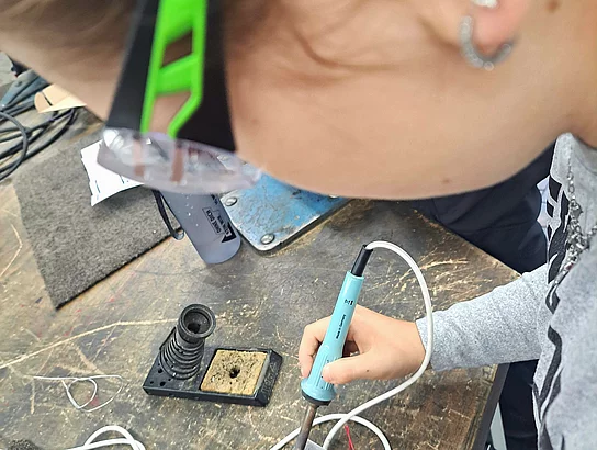Nahaufnahme einer Person mit Schutzbrille, die an einer Werkbank mit einem elektrischen Lötkolben an einem kleinen Bauteil arbeitet.