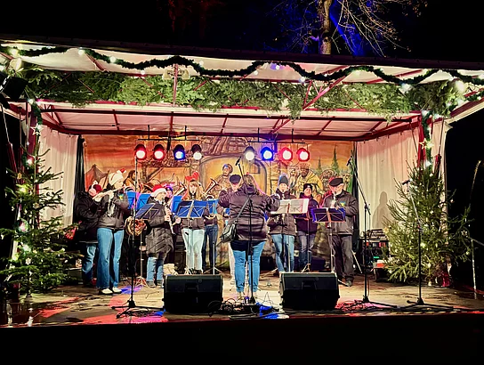 Zu sehen ist ein Blasmusikensemble auf einer festlich beleuchteten Bühne bei Einbruch der Dunkelheit in einem Stadtpark.