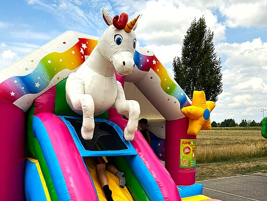 Ein großes aufblasbares Einhorn ziert eine farbenfrohe Hüpfburg, auf der Kinder spielen und rutschen.