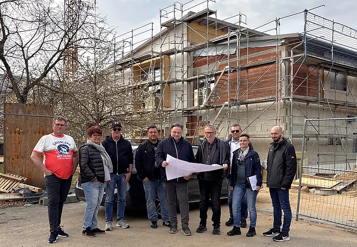 Mehrere Personen stehen vor einem Gebäude im Bau mit Gerüst und Baukran und betrachten gemeinsam einen ausgebreiteten Bauplan.