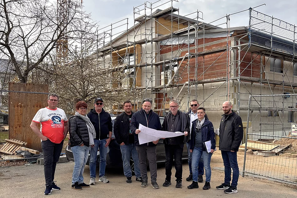 Mehrere Personen stehen vor einem Gebäude im Bau mit Gerüst und Baukran und betrachten gemeinsam einen ausgebreiteten Bauplan.