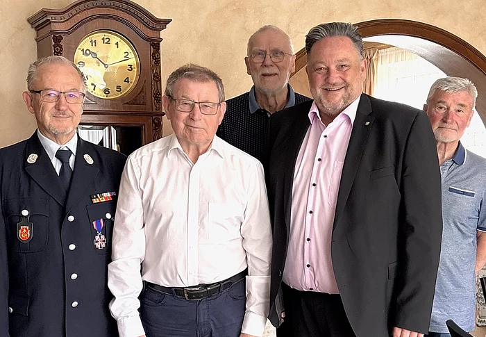 Fünf Männer stehen gemeinsam in einem Wohnraum und blicken in die Kamera. Einer der Männer trägt eine Feuerwehruniform mit Auszeichnungen, die anderen sind in Hemd, Sakko oder Poloshirt gekleidet. In der Mitte steht ein älterer Herr im weißen Hemd. Im Hintergrund sind eine große Standuhr sowie ein Rundbogen zu sehen. Die Aufnahme vermittelt den Eindruck eines offiziellen Geburtstags- oder Ehrenbesuchs in persönlicher Atmosphäre.