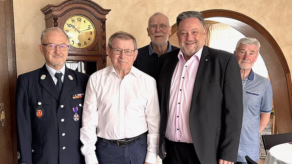 Fünf Männer stehen gemeinsam in einem Wohnraum und blicken in die Kamera. Einer der Männer trägt eine Feuerwehruniform mit Auszeichnungen, die anderen sind in Hemd, Sakko oder Poloshirt gekleidet. In der Mitte steht ein älterer Herr im weißen Hemd. Im Hintergrund sind eine große Standuhr sowie ein Rundbogen zu sehen. Die Aufnahme vermittelt den Eindruck eines offiziellen Geburtstags- oder Ehrenbesuchs in persönlicher Atmosphäre.