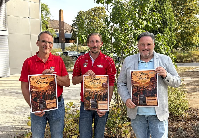 Zwei Vertreter der FG D’Rummlfelser und Bürgermeister Thomas Beer stehen vor der Dreifachturnhalle und halten jeweils ein Plakat für die Veranstaltung „Tanz in den Herbst“ in den Händen.