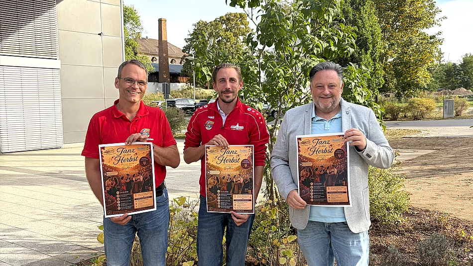 Zwei Vertreter der FG D’Rummlfelser und Bürgermeister Thomas Beer stehen vor der Dreifachturnhalle und halten jeweils ein Plakat für die Veranstaltung „Tanz in den Herbst“ in den Händen.