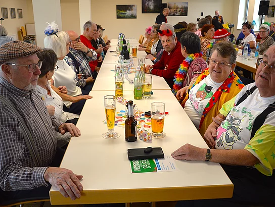 Zwei Reihen älterer Menschen sitzen sich an einem langen Tisch gegenüber, trinken Bier oder Wasser und tragen fröhliche, teilweise sehr bunte Faschingskleidung.