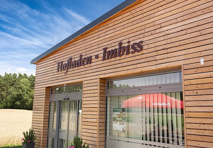 Das Foto zeigt den modernen Holzbau des Hofladens und Imbisses des Teichguts Mulzer mit großen Glasfronten und der gut sichtbaren Aufschrift „Hofladen · Imbiss“. Im Hintergrund sind ein Getreidefeld und ein Waldrand unter blauem Himmel zu erkennen.
