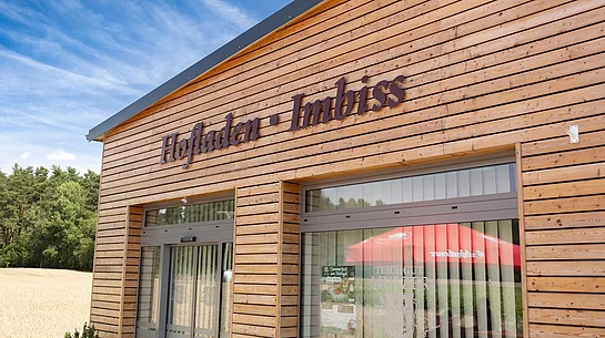 Das Foto zeigt den modernen Holzbau des Hofladens und Imbisses des Teichguts Mulzer mit großen Glasfronten und der gut sichtbaren Aufschrift „Hofladen · Imbiss“. Im Hintergrund sind ein Getreidefeld und ein Waldrand unter blauem Himmel zu erkennen.