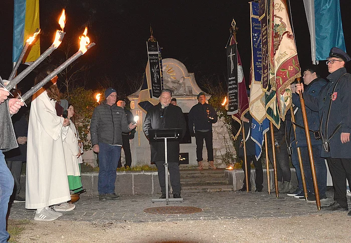 Eine Gruppe von Menschen steht abends in einem Halbkreis mit brennenden Fackeln und Fahnen um einen Redner vor einem beleuchteten Denkmal.