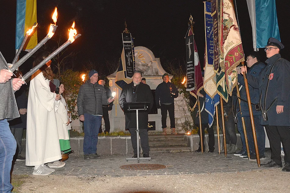 Eine Gruppe von Menschen steht abends in einem Halbkreis mit brennenden Fackeln und Fahnen um einen Redner vor einem beleuchteten Denkmal.