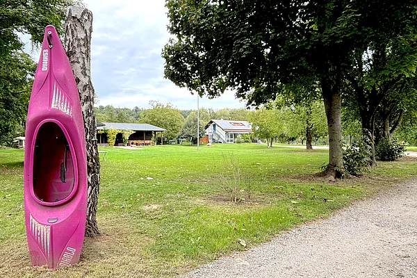 Blick über die Liegewiese in Richtung zweier flacher Bungalow-Gebäude. Im Vordergrund ein an einen Baumstumpf gelehnte pinkes Kajak.