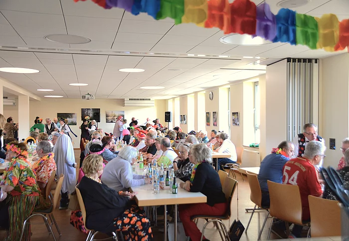 In einem Saal sitzen an langen Tischen zahlreiche bunt maskierte Seniorinnen und Senioren und im Hintergrund sind man einige Tanzpaare. 