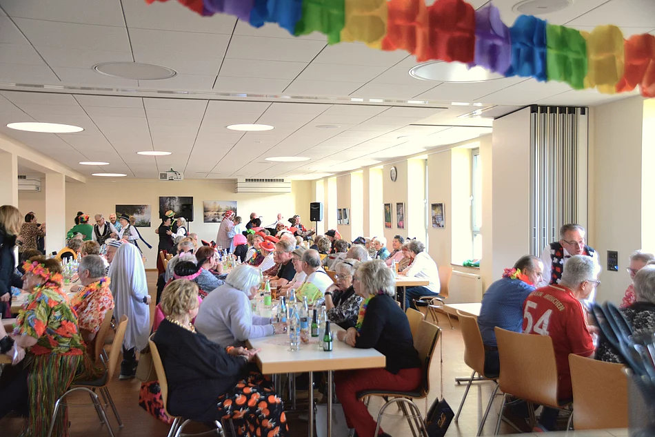In einem Saal sitzen an langen Tischen zahlreiche bunt maskierte Seniorinnen und Senioren und im Hintergrund sind man einige Tanzpaare. 