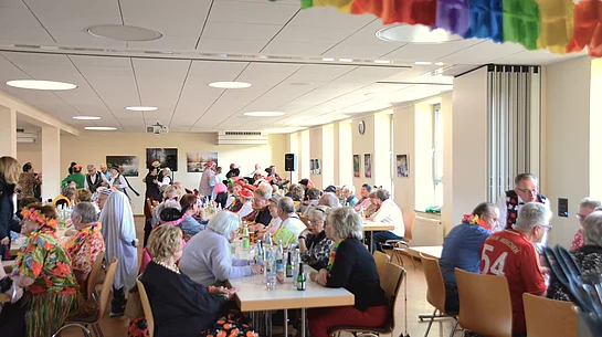 In einem Saal sitzen an langen Tischen zahlreiche bunt maskierte Seniorinnen und Senioren und im Hintergrund sind man einige Tanzpaare. 