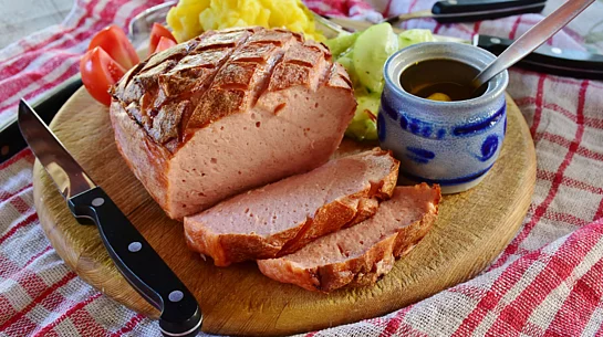 Auf einem Holzbrett liegt ein aufgeschnittener Leberkäse mit knuspriger Kruste, daneben Kartoffelpüree, Gurkensalat, Senf und ein Messer, angerichtet wie ein traditionelles bayerisches Gericht.