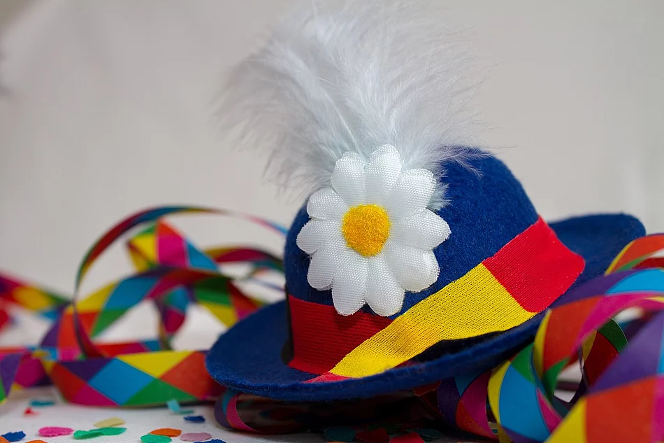 Das Bild zeigt einen traditionellen Faschingshut in Blau mit bunter Schleife, weißer Feder und Blume, umgeben von farbigem Konfetti und Luftschlangen als Symbol für ausgelassene Karnevalsstimmung.