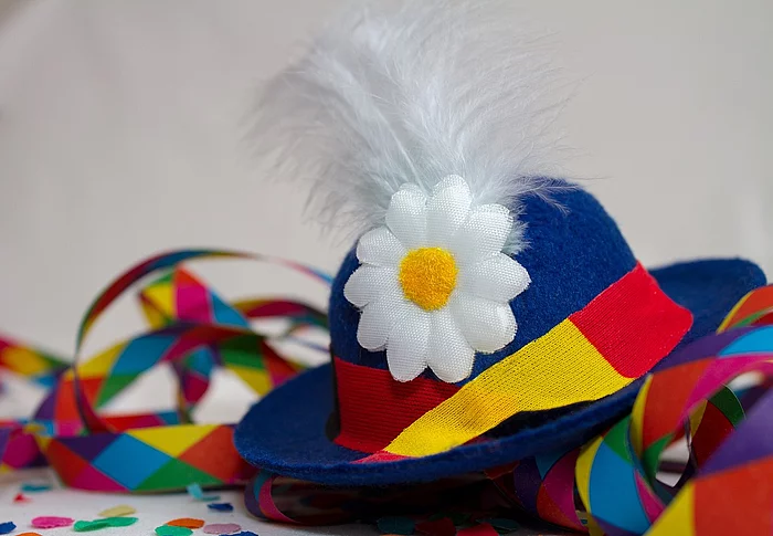 Das Bild zeigt einen traditionellen Faschingshut in Blau mit bunter Schleife, weißer Feder und Blume, umgeben von farbigem Konfetti und Luftschlangen als Symbol für ausgelassene Karnevalsstimmung.