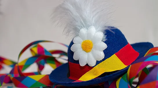 Das Bild zeigt einen traditionellen Faschingshut in Blau mit bunter Schleife, weißer Feder und Blume, umgeben von farbigem Konfetti und Luftschlangen als Symbol für ausgelassene Karnevalsstimmung.