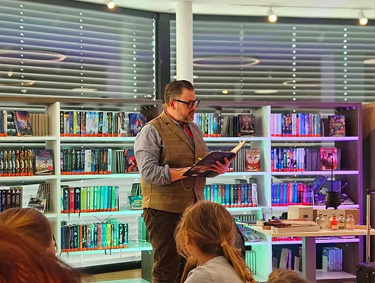 Ein Mann steht vor einem Publikum aus Kindern und Erwachsenen und liest aus einem Buch vor. Hinter ihm befinden sich bunt beleuchtete Bücherregale. Die Zuhörer sitzen aufmerksam vor der kleinen Lesebühne in der Bücherei.