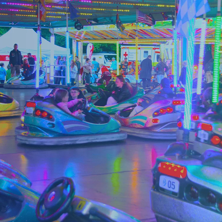 Mehrere Menschen fahren in bunten Autoscootern auf einem beleuchteten Volksfest-Fahrgeschäft, während im Hintergrund Besucher zwischen Zelten und Ständen stehen.