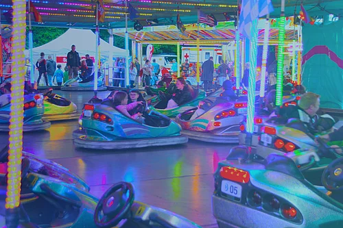 Mehrere Menschen fahren in bunten Autoscootern auf einem beleuchteten Volksfest-Fahrgeschäft, während im Hintergrund Besucher zwischen Zelten und Ständen stehen.