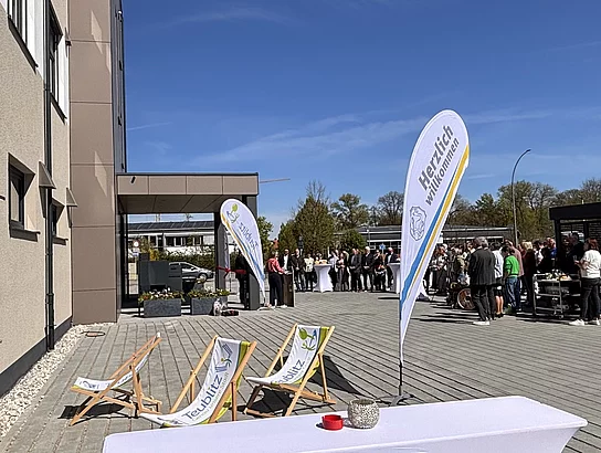 Blick auf den Veranstaltungsbereich vor dem Gebäude mit Gästen und Fahnen zur Feier der Neueröffnung.