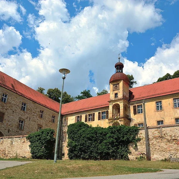 Ein Renaissance-Schloss mit Steinbauern und mehreren Gebäudeflügeln. In der Mitte ein Glockenturm. Das Gebäude thront auf einem grünen Hügel.