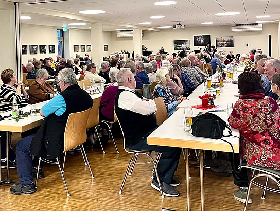 Ein großer Saal mit mehreren Tischreihen, die jeweils komplett voll besetzt sind. 