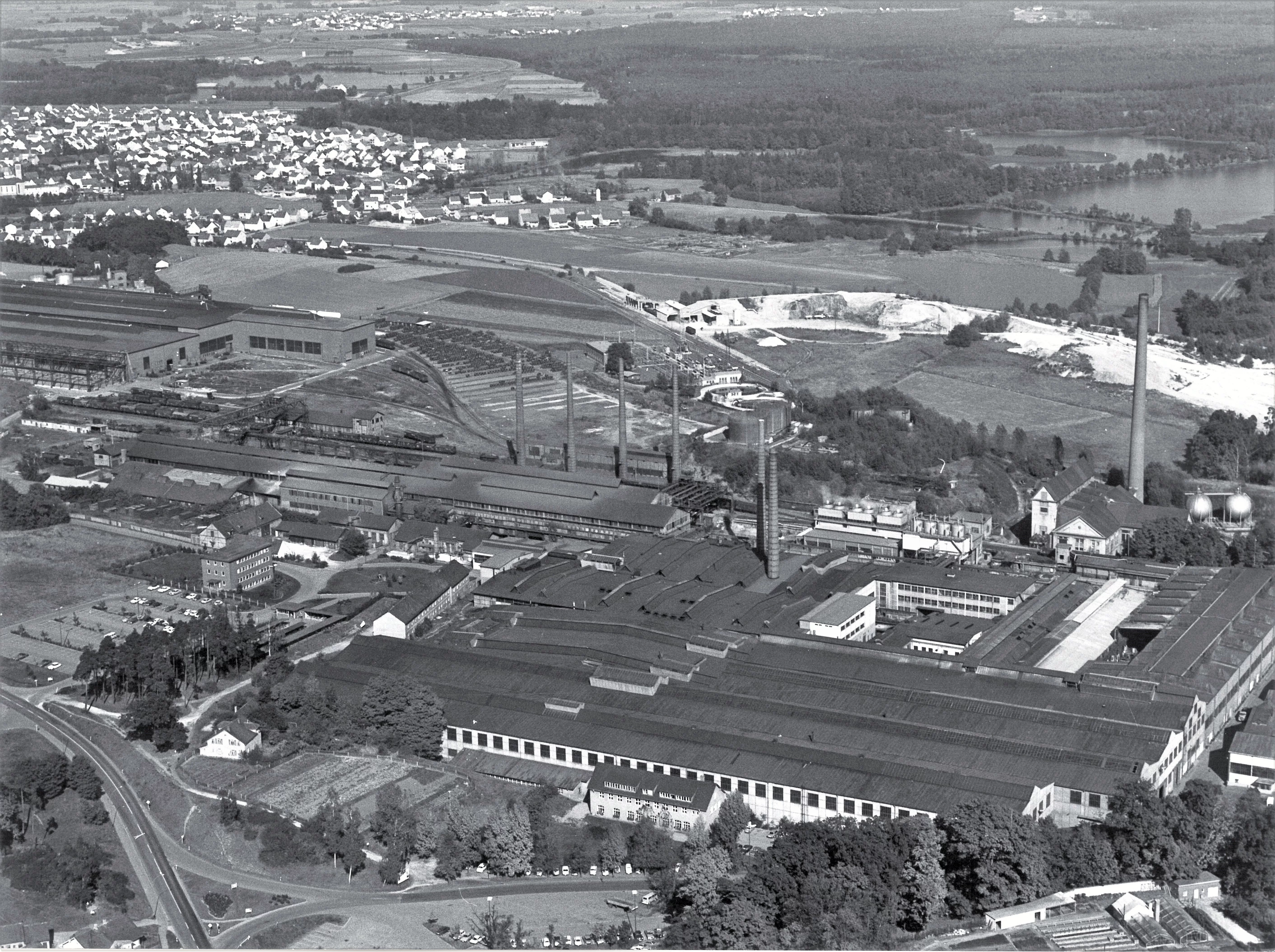 Eine Luftbildaufnahme in schwarz-weiß von einer sehr großen Industriellen Hallenanlage mit großem Schornstein.