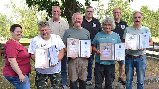 Eine Gruppe von acht Personen steht im Freien vor Bäumen und einem Holzzaun. Vier langjährige Urlaubsgäste halten Ehrenurkunden und Dankesurkunden der Stadt Teublitz in den Händen. Hinter ihnen stehen Vertreter der Stadt, des Fischereivereins und des Landgasthofs Hintermeier. Die Geehrten lächeln in die Kamera und präsentieren ihre Auszeichnungen. Das Foto entstand im Rahmen der Ehrung für langjährige Urlaubstreue.