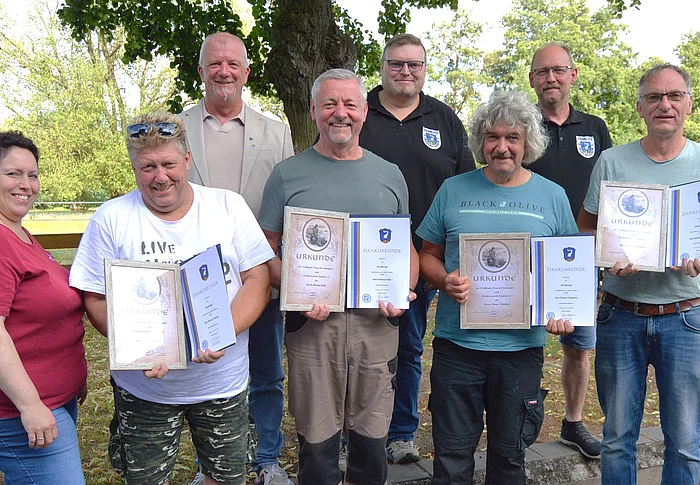 Eine Gruppe von acht Personen steht im Freien vor Bäumen und einem Holzzaun. Vier langjährige Urlaubsgäste halten Ehrenurkunden und Dankesurkunden der Stadt Teublitz in den Händen. Hinter ihnen stehen Vertreter der Stadt, des Fischereivereins und des Landgasthofs Hintermeier. Die Geehrten lächeln in die Kamera und präsentieren ihre Auszeichnungen. Das Foto entstand im Rahmen der Ehrung für langjährige Urlaubstreue.