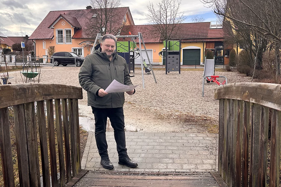 Ein Mann steht auf einer kleinen Holzbrücke vor einem Spielplatz und hält ein Blatt Papier in der Hand, während hinter ihm Spielgeräte und Wohnhäuser zu sehen sind.