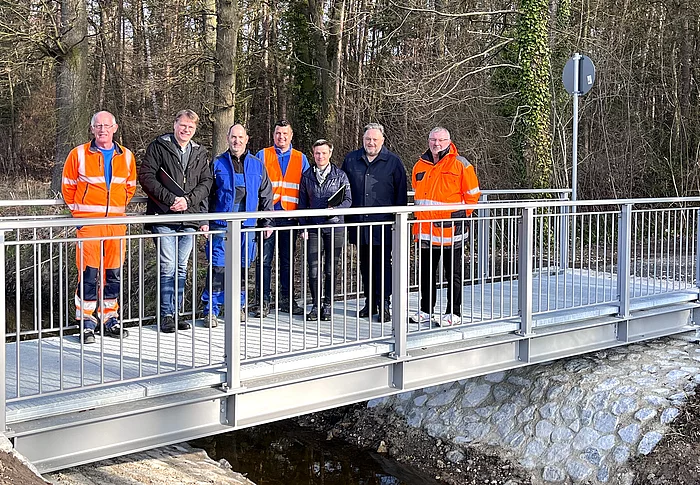Eine Gruppe von Personen steht gemeinsam auf einer neu errichteten kleinen Brücke über einen Bach vor einem bewaldeten Hintergrund.