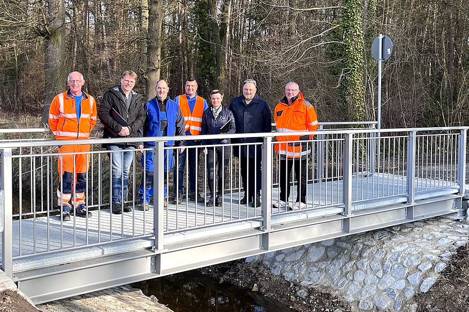 Eine Gruppe von Personen steht gemeinsam auf einer neu errichteten kleinen Brücke über einen Bach vor einem bewaldeten Hintergrund.