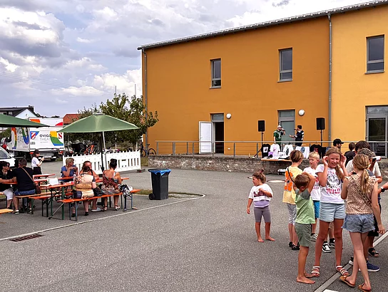Kinder und Familien verfolgen das Bühnenprogramm vor dem Mehrgenerationenhaus, während andere an den Bierbänken zusammensitzen.
