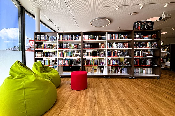 Heller Bibliotheksraum mit großen Fensterfronten und mehreren Bücherregalen voller Romane und Fantasy-Literatur. Im Vordergrund stehen grüne Sitzsäcke und ein roter Hocker als gemütliche Leseplätze. Die moderne Gestaltung schafft eine entspannte Atmosphäre zum Lesen und Entdecken.
