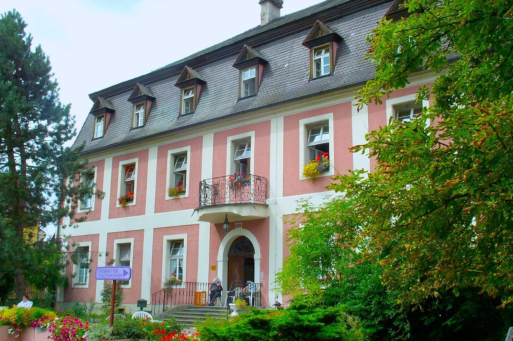 Blick auf ein prächtiges Gebäude mit Mansarddach und zartrosa Farbe, vielen Fenstern, abgerundeter Flügeltür. Im Vordergrund ein sehr großer Nadelbaum.