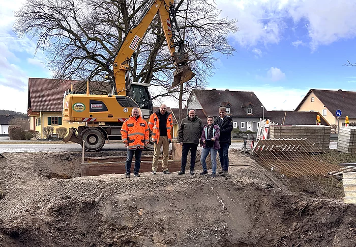 Vor einem großen Bagger stehen mehrere Personen am Rand einer ausgehobenen Baugrube, in der ein Brückenbau vorbereitet wird.